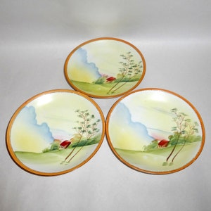 May include: Three white ceramic plates with a hand-painted design of a red house, green trees, and blue clouds on a yellow background. The plates have a brown rim.