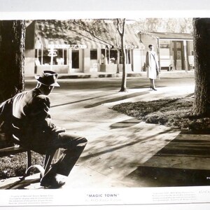 May include: A black and white photograph of a man wearing a hat sitting on a bench in front of a building. The building has a sign that reads "Magic Town".