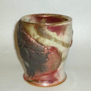 May include: A ceramic cup featuring a textured, dark brown, cracked-earth exterior panel contrasted against a smooth, glossy glaze in shades of cream, muted green, and deep red. The vessel has a wide, open rim and a slightly flared base.