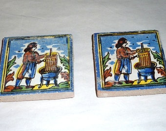 May include: Two square ceramic tiles with a hand-painted scene of a person operating a press. The tiles have a blue border, a blue sky background, and colorful details. The person is wearing a brown jacket and blue pants.