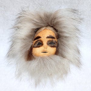 May include: A handmade mask with a human face, crafted from leather and fur. The mask has a long, white fur beard and a white fur hood. The eyes are black and the face is painted with brown details.