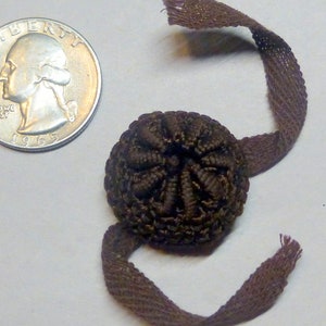 May include: A brown fabric button with a decorative design and a long brown fabric loop attached.