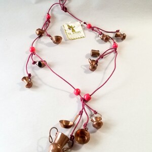 May include: A necklace featuring miniature copper kitchenware charms, including pots, pitchers, and ladles, strung on a maroon cord with red beads. The charms are detailed and shiny. A tag with the text "EL ANGEL" is attached.