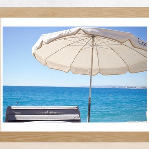 Può includere: Un ombrellone da spiaggia bianco con la scritta "Letaba" stampata su di esso, che proietta ombra su una sedia a sdraio grigia. L'ombrellone è posizionato su una spiaggia sabbiosa con vista sull'oceano blu e una costa lontana.