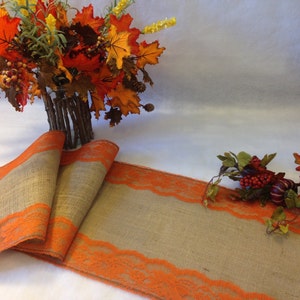 May include: A burlap table runner with orange lace trim. The runner is folded in half and is lying on a white surface.