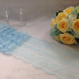 May include: A light blue lace ribbon with a delicate floral pattern. The ribbon is folded and displayed on a white surface. A bouquet of yellow roses and blue hydrangeas is in the background.