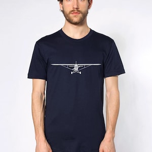 May include: Navy blue t-shirt featuring a white line drawing of a small airplane. The graphic is centered on the chest. The shirt has a crew neck and short sleeves. The person in the image is wearing dark blue jeans.