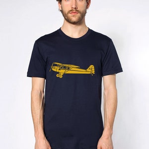 Puede incluir: Una camiseta azul marino con una silueta amarilla de un avión vintage impresa en la parte delantera.