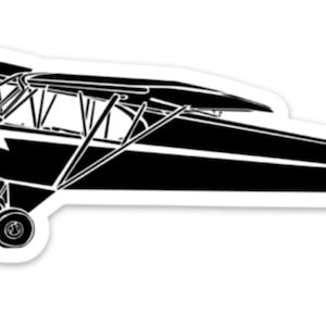 Könnte beinhalten: Schwarz-weiße Silhouette eines kleinen Flugzeugs mit einem einzelnen Propeller. Das Flugzeug ist im Flug und hat eine sichtbare Kennung.