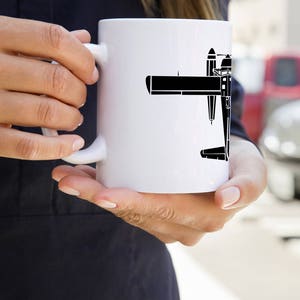 May include: A white ceramic mug with a black silhouette of a seaplane on it. The mug is being held by a person's hand.