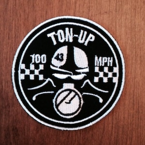 May include: Black and white embroidered patch with the text "TON-UP" and "100 MPH". The patch features a cartoon illustration of a person wearing a helmet and riding a motorcycle.