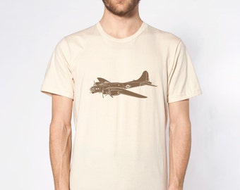 Puede incluir: Un hombre con una camiseta de color crema con un avión vintage de color marrón impreso en la parte delantera. El hombre lleva pantalones vaqueros azules.