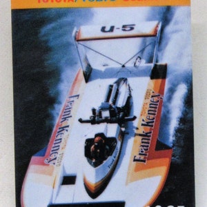 May include: A white and orange hydroplane boat with the text "Frank Kenney" and "TOYOTA VOLVO" on the side. The boat is in the air, creating a spray of water. The text "U-5" is visible on the top of the boat. The year "1985" is printed in white on a black background at the bottom of the image.