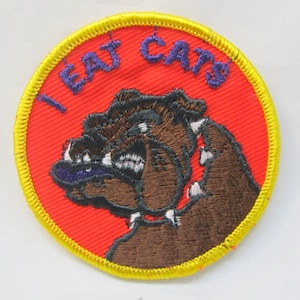 Può includere: Una toppa ricamata rossa e gialla con un bulldog di cartone animato che indossa un collare a punte. Il testo "I EAT CATS" è ricamato in viola intorno al bulldog.