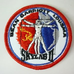 Puede incluir: Un parche bordado blanco, rojo y azul con el texto "Bean Garriott Louma Skylab II". El parche presenta una imagen estilizada de una persona con los brazos extendidos.