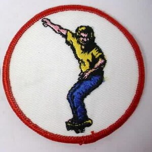 Puede incluir: Un parche bordado blanco con borde rojo, que presenta una ilustración de dibujos animados de una persona haciendo skateboard con una camisa amarilla y pantalones vaqueros azules.