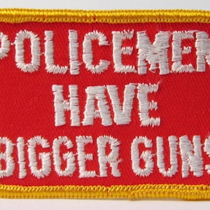 Pode incluir: Um remendo vermelho bordado com uma borda amarela. O texto no remendo diz "Policemen Have Bigger Guns".