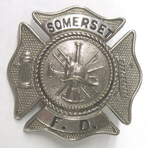 May include: A silver firefighter's badge with the word "SOMERSET" at the top. The centre features a helmet, axes, and other tools. The bottom reads "F.D."