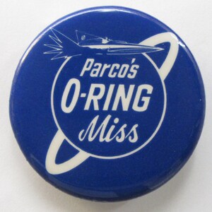 Op de afbeelding: Een blauwe knop met een witte afbeelding van een raket die door een ring vliegt. De tekst "Parco's O-RING Miss" staat op de knop gedrukt.