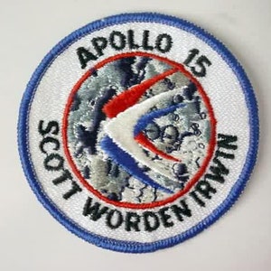 Może przedstawiać: Biała naszywka haftowana z niebieskim obramowaniem. Naszywka przedstawia czerwony, biały i niebieski wzór z tekstem "Apollo 15" i "Scott Worden Irwin".