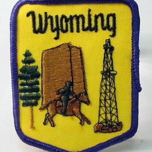 Op de afbeelding: Gele geborduurde patch met paarse rand met het woord "Wyoming" in zwarte letters. De patch toont een bruin gebouw, een groene dennenboom, een zwart-witte olieboringstoren en een bruin paard met een ruiter.
