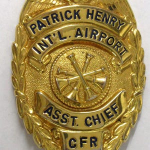 May include: A gold-colored badge with the text "PATRICK HENRY INT'L. AIRPORT" and "ASST. CHIEF CFR" in blue lettering. The badge features a central emblem with crossed bugles and is surrounded by a laurel wreath design.