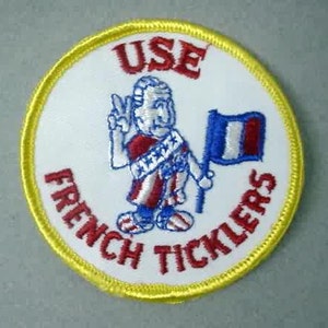 Könnte beinhalten: Ein weißer gestickter Aufnäher mit gelbem Rand. Der Aufnäher zeigt eine Comicfigur, die eine Flagge mit den Farben der französischen Flagge hält. Der Text "USE FRENCH TICKLERS" ist in Rot um die Figur herum gestickt.
