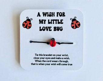 May include: A white card with black text that reads "A Wish For My Little Love Bug". Two red and black ladybug illustrations are on either side of the text. A black string bracelet with a red and black ladybug charm is on the card.