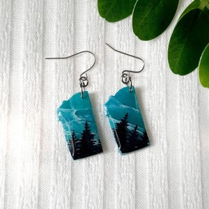 K&ouml;nnte beinhalten: Ein Paar silberne Ohrh&auml;nger mit einem blauen und schwarzen Harzdesign, das eine Silhouette eines Waldes zeigt.