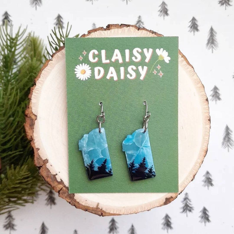 K&ouml;nnte beinhalten: Ein Paar H&auml;ngeohrringe mit einem blauen und schwarzen Design, das an Achat erinnert. Die Ohrringe befinden sich auf einer gr&uuml;nen Karte mit dem Text "Claisy Daisy" in Wei&szlig;.