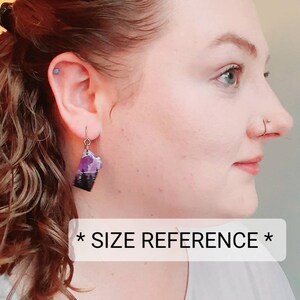 K&ouml;nnte beinhalten: Eine Person tr&auml;gt ein wei&szlig;es Shirt mit dem Text "* SIZE REFERENCE *" und tr&auml;gt ein Paar H&auml;ngeohrringe mit einem lila und schwarzen Design.