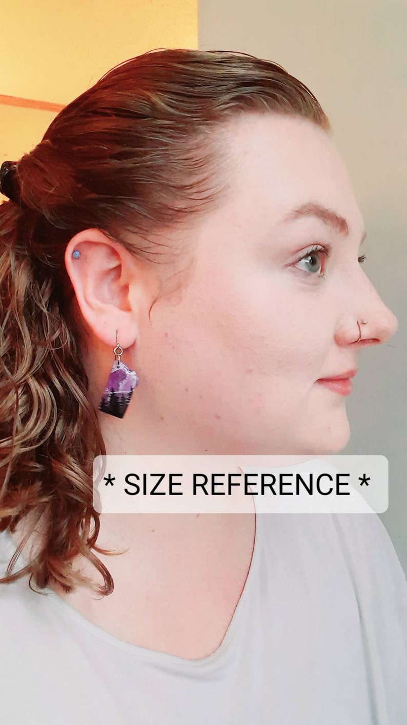 K&ouml;nnte beinhalten: Eine Person tr&auml;gt ein wei&szlig;es Shirt mit dem Text "* SIZE REFERENCE *" und tr&auml;gt ein Paar H&auml;ngeohrringe mit einem lila und schwarzen Design.