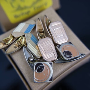 Op de afbeelding: Een verzameling vintage manchetknopen in diverse geometrische ontwerpen. De manchetknopen hebben gouden, zilveren en emaille accenten in kleuren als blauw, wit, zwart en oranje. Sommige hebben streepjespatronen, andere getextureerde oppervlakken.