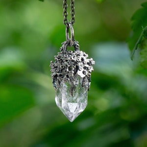 May include: A silver pendant necklace with a clear crystal point encased in a silver floral design. The necklace is hanging from a branch with green leaves.
