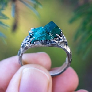 May include: A silver ring with a green gemstone set in a leafy design. The ring is on a finger against a blurred background of green foliage.