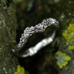 May include: A silver ring with a textured, organic surface resembling tree bark and small, raised circular nodes along the top edge. The metal has a dark, oxidised finish in the recessed areas, highlighting the intricate, uneven details of the band.