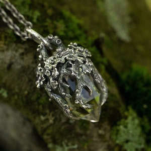 May include: A silver necklace with a clear quartz crystal pendant encased in a detailed silver cage. The pendant is hanging from a silver chain against a mossy green background.