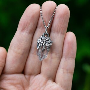 May include: A silver necklace with a clear quartz crystal pendant. The pendant is surrounded by a delicate silver design that resembles branches or vines.