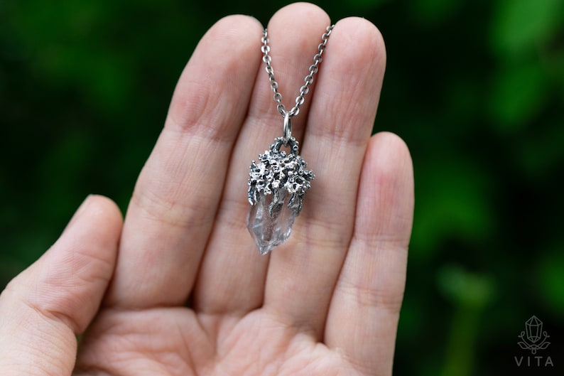 May include: A silver necklace with a clear quartz crystal pendant. The pendant is surrounded by a delicate silver design that resembles branches or vines.
