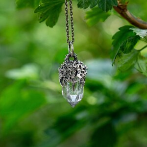 May include: A silver necklace with a clear quartz crystal pendant encased in a silver, tree-like design. The necklace is hanging from a branch with green leaves.