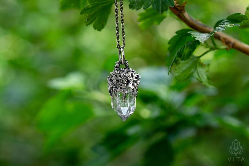 May include: A silver necklace with a clear quartz crystal pendant encased in a silver, tree-like design. The necklace is hanging from a branch with green leaves.