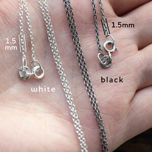 May include: Two silver chains, one white and one black, both 1.5mm thick. The chains are laid out on a hand, with the clasps visible. The chains are both made of sterling silver.