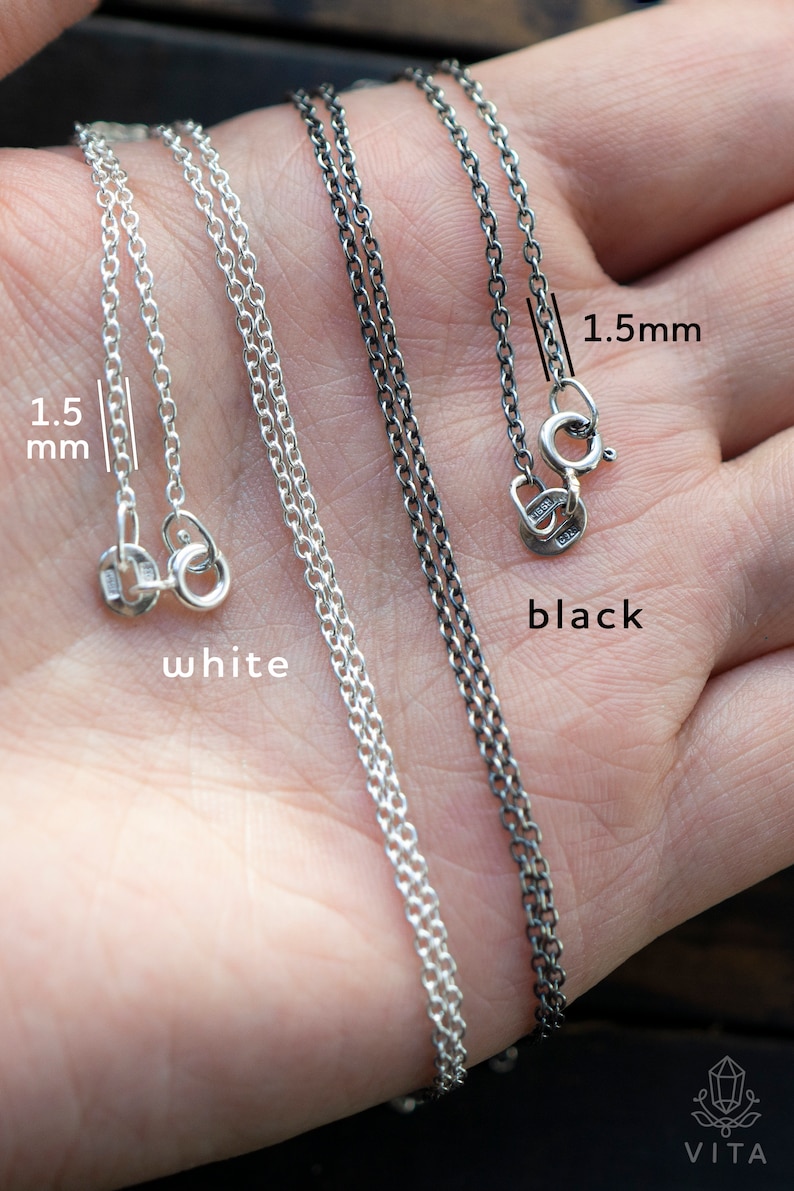 May include: Two silver chains, one white and one black, both 1.5mm thick. The chains are laid out on a hand, with the clasps visible. The chains are both made of sterling silver.