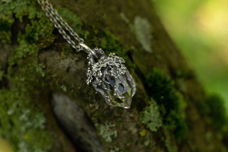 May include: A silver necklace with a clear quartz crystal pendant encased in a detailed silver cage. The pendant is hanging from a silver chain against a mossy green background.