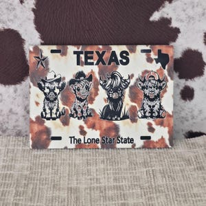 May include: A rectangular license plate featuring a cowhide pattern background with the word "TEXAS" at the top. The plate has four cartoon Highland cows wearing hats. The text "The Lone Star State" is at the bottom.