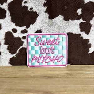 May include: A rectangular patch with a checkered turquoise and white pattern, framed in pink. The patch features the embroidered text "Sweet but PSYCHO" in pink script. The background is a cowhide pattern.