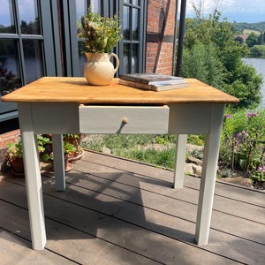 Könnte beinhalten: Ein rechteckiger Holztisch mit einer naturfarbenen, hellbraunen Holzmaserung auf der Tischplatte und einem hellgrau lackierten Untergestell. Der Tisch hat eine mittige Schublade mit einem kleinen runden Holzknopf. Ein cremefarbener Krug mit Blumen und zwei Bücher stehen darauf.