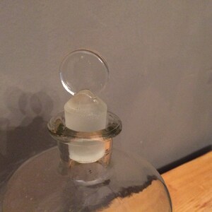 May include: A clear glass bottle with a glass stopper. The stopper has a round, flat top.