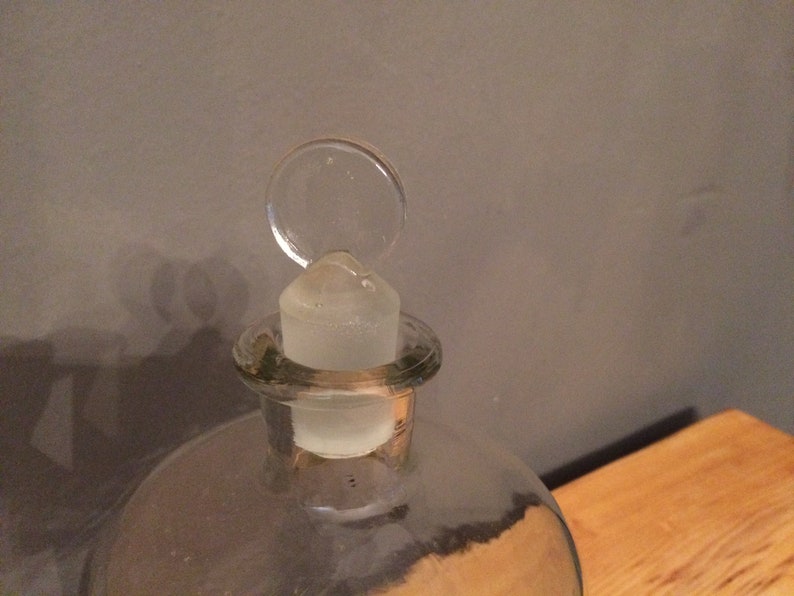 May include: A clear glass bottle with a glass stopper. The stopper has a round, flat top.