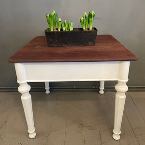 Könnte beinhalten: Ein weißer Tisch mit einer dunkelbraunen Holzplatte. Der Tisch hat vier gedrehte Beine und ein schlichtes Design. Ein Topf mit grünen Hyazinthenpflanzen steht auf dem Tisch.
